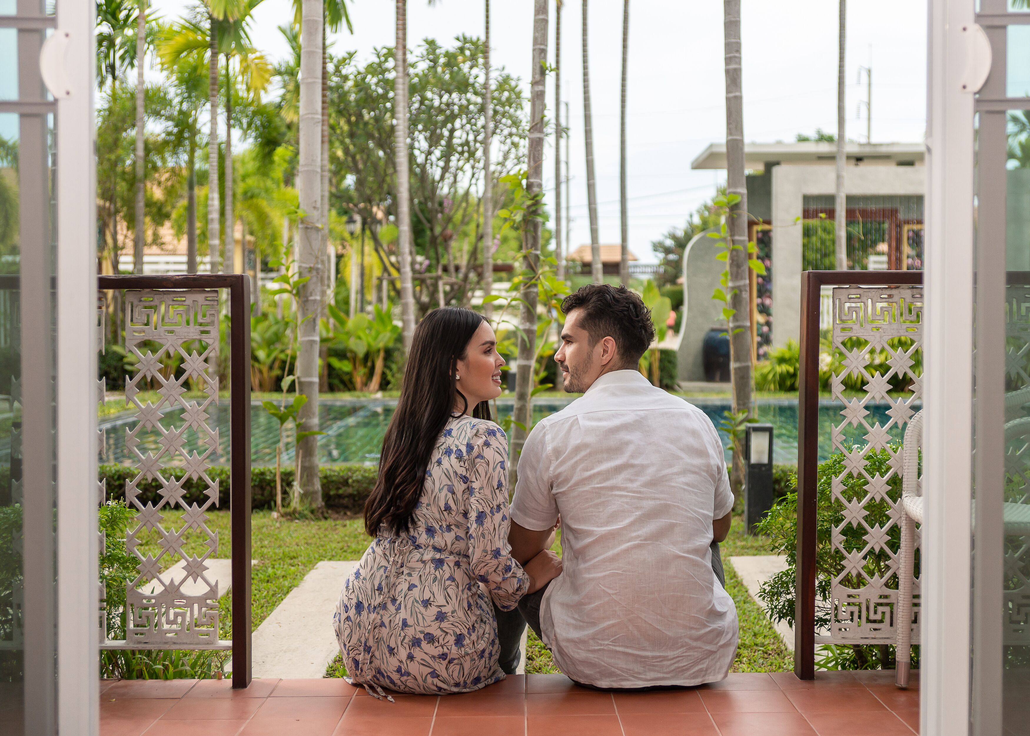 A couple sitting on a tiled patio with a decorative gate and lush greenery in the background.