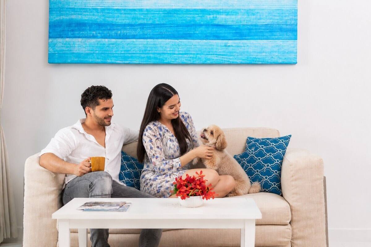 Ein Paar sitzt auf einem beigefarbenen Sofa mit einem blau gemusterten Kissen, einem kleinen Tisch mit einer Zeitschrift und einer roten Blumenvase.