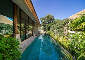 Une piscine dont les bords sont recouverts de carreaux, entourée d’une végétation luxuriante et bordée d’un bâtiment doté de grandes fenêtres.