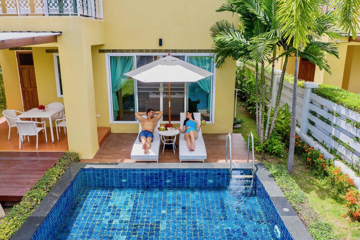 Ein Schwimmbecken mit einer gefliesten Kante, eine Terrasse mit Tisch und Stühlen und Blick auf ein Gebäude mit großen Fenstern und einer Tür.