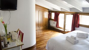 Et hotelværelse med seng, fjernsyn ophængt på væggen, trægulv, et vindue med gardiner og et lille bord med en vase og et glas.
