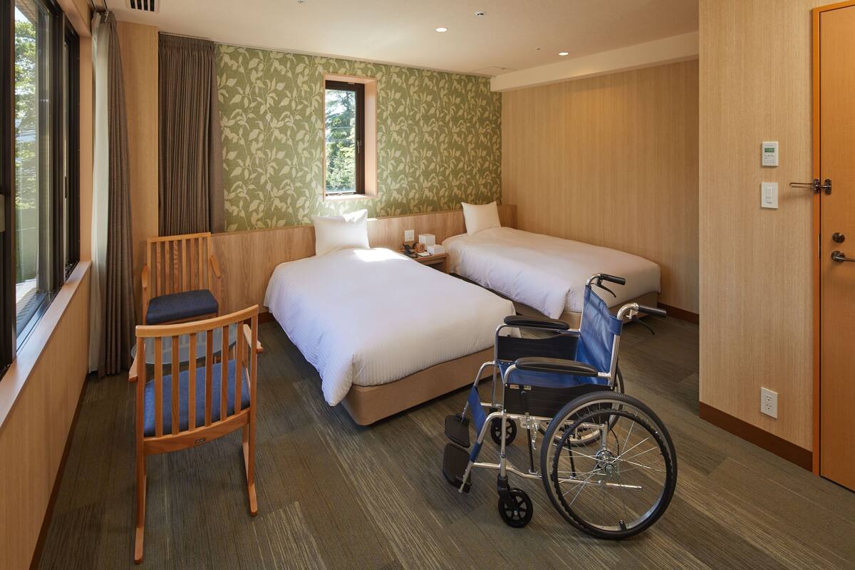 A hotel room with two beds, a wheelchair, a chair, and a window.