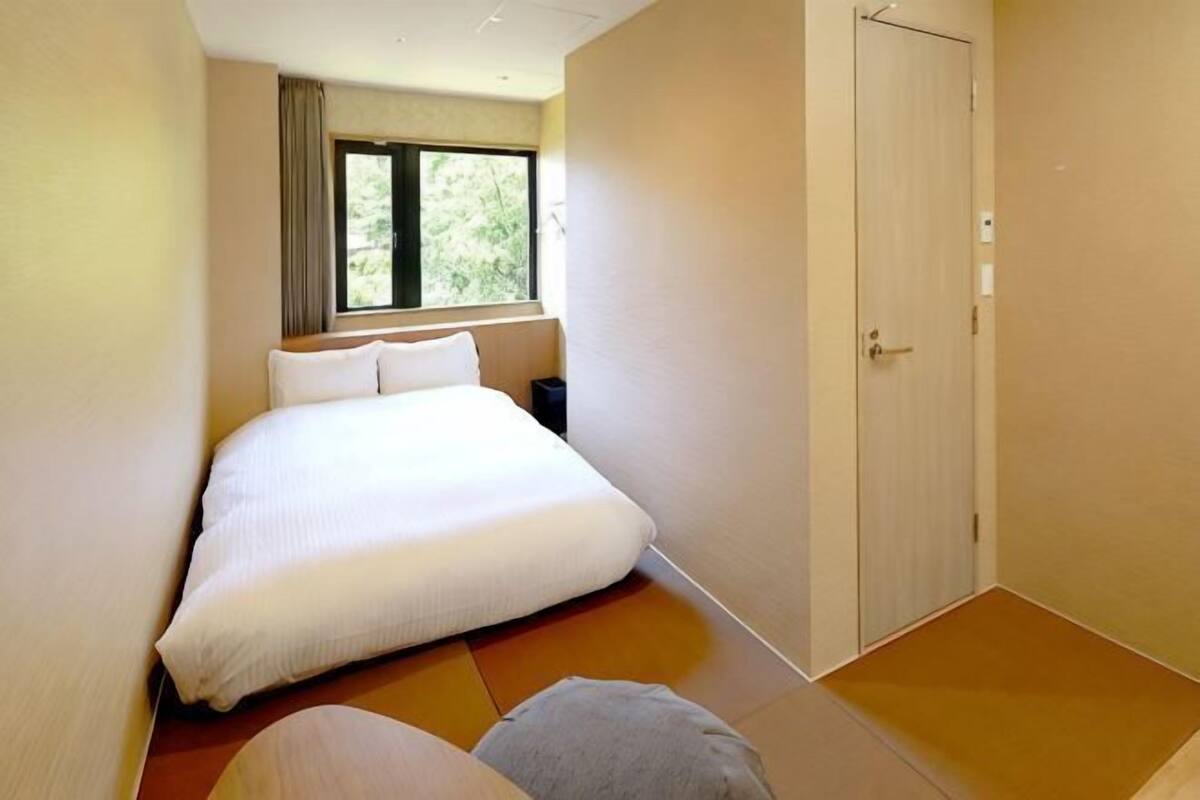 A hotel room with a bed, a window, a door, and a small bedside table.