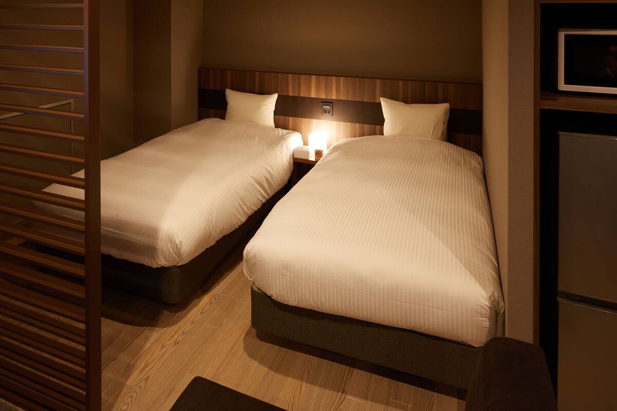 A hotel room with two single beds, a nightstand with a lamp, a wooden headboard, and a small refrigerator.