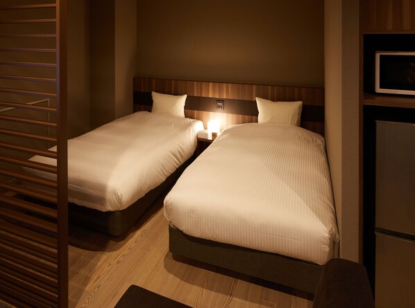 A hotel room with two single beds, a nightstand with a lamp, a wooden headboard, and a small refrigerator.