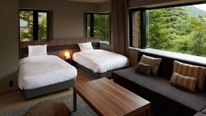 A hotel room with two beds, a sofa, a wooden table, and large windows offering a view of greenery. - Hakone Yutowa (Hakone)