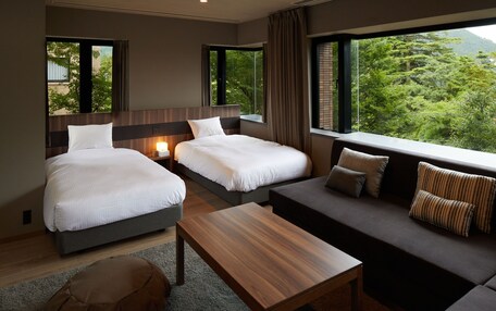 A hotel room with two beds, a sofa, a wooden table, and large windows offering a view of greenery.