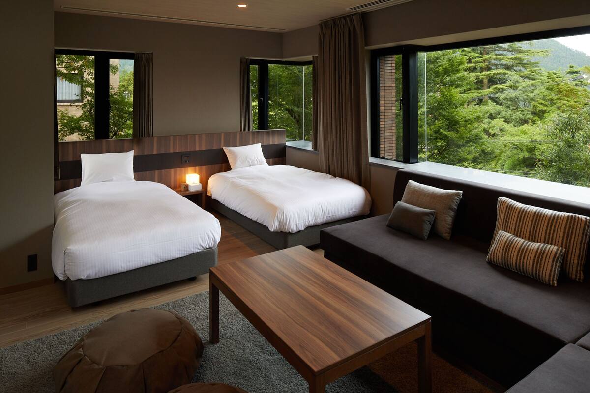 A hotel room with two beds, a sofa, a wooden table, and large windows offering a view of greenery.