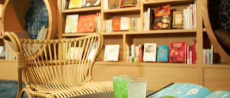 A library with bookshelves, a wicker chair, and a table with a book and glasses of water.