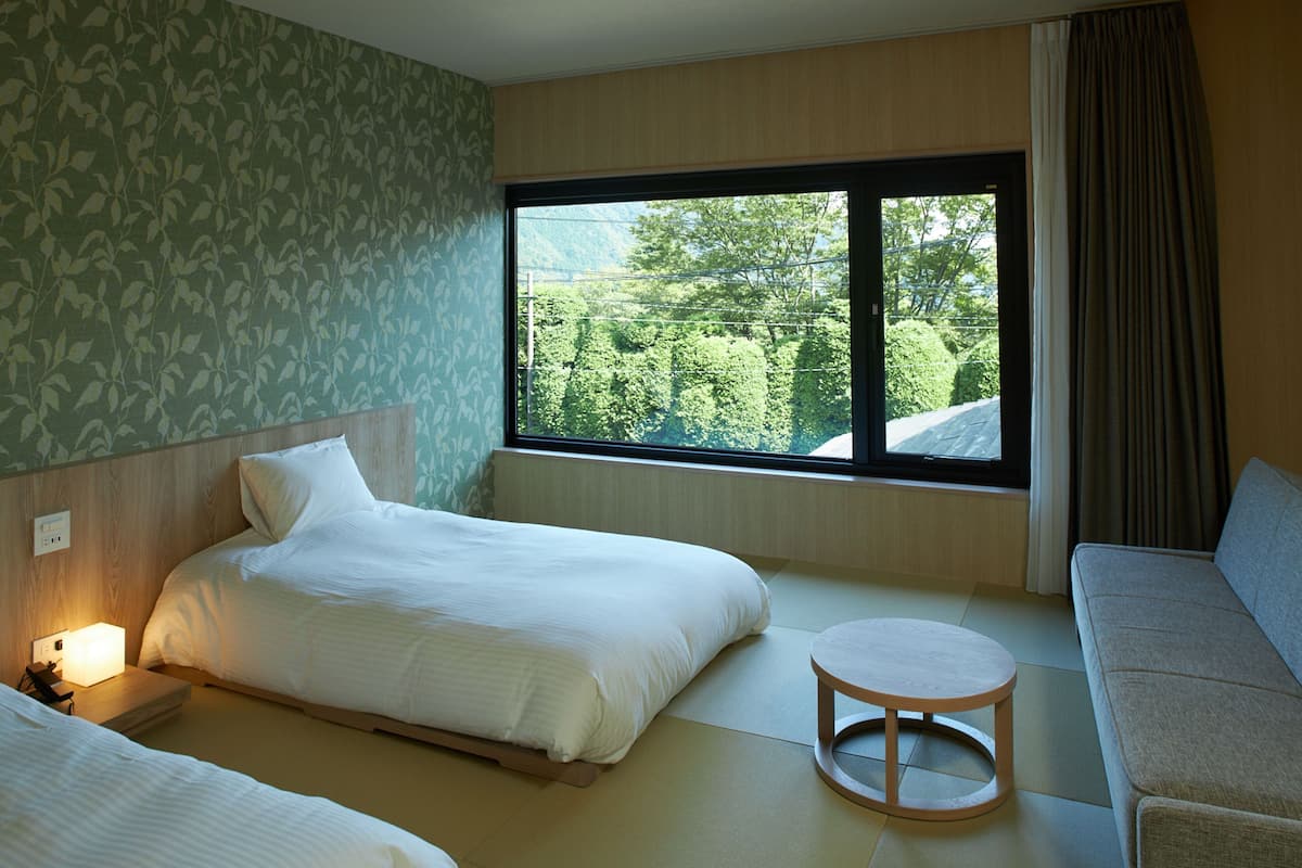 A hotel room with two beds, a sofa, a small round table, and a large window offering a view of greenery.