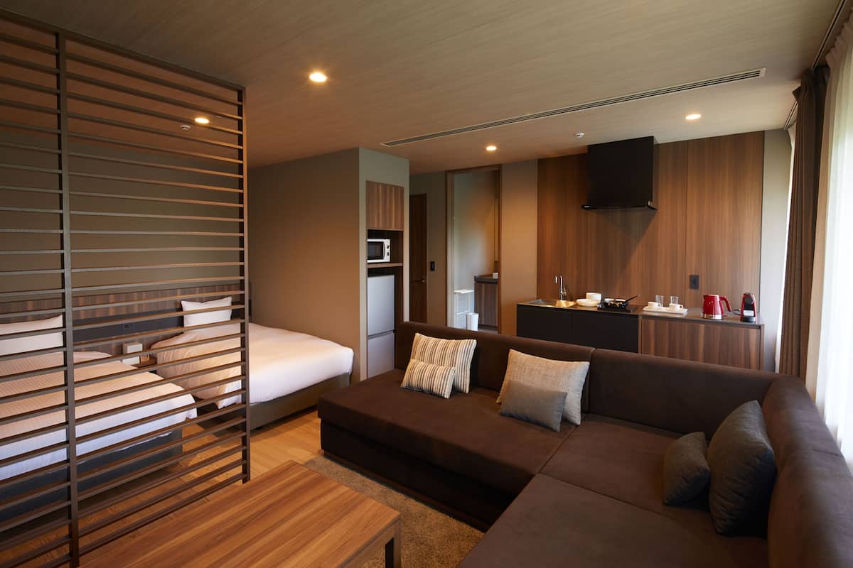 A modern hotel room with a bed, a sofa, a wooden table, a kitchenette, and a TV mounted on the wall.