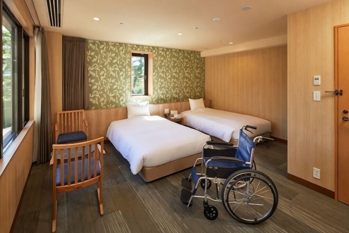 A hotel room with two beds, a wheelchair, a wooden chair, and a window.
