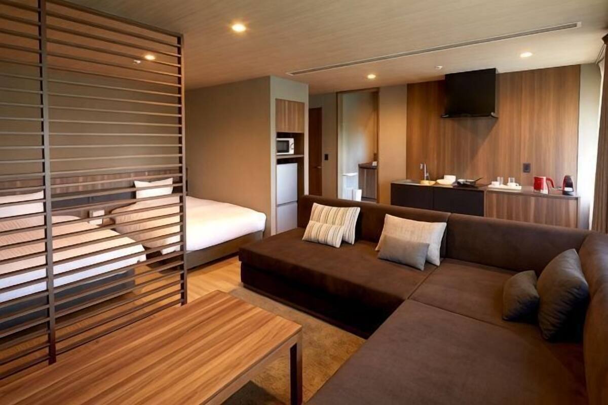 A modern hotel room with a sofa, a wooden coffee table, a bed with a headboard, a kitchenette, and a TV mounted on the wall.