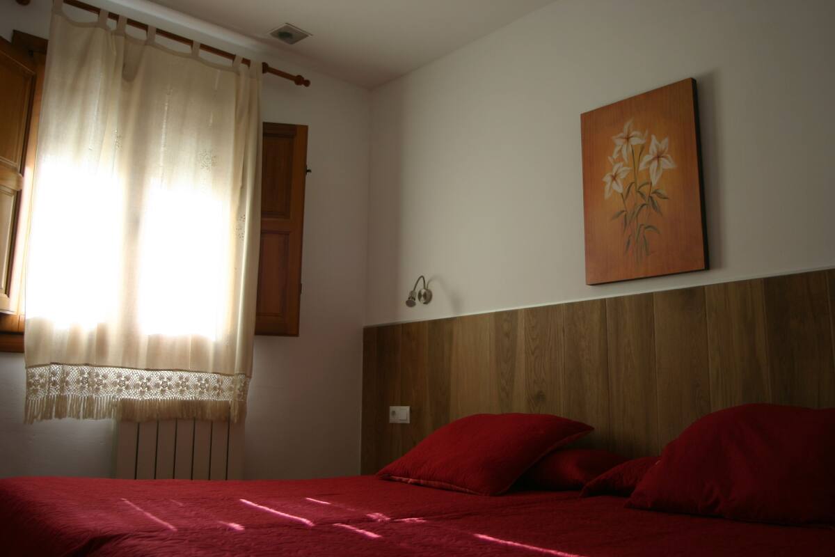 Een slaapkamer met een houten hoofdbord, een rode sprei en een bloemen schilderij aan de muur.
