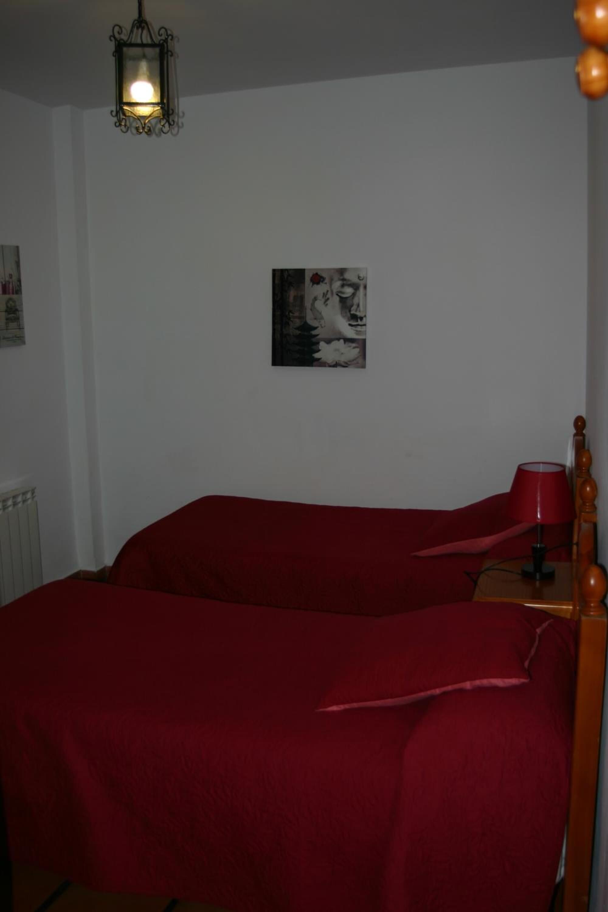 Een kamer met een bed met rode beddengoed, een houten nachtkastje met een rode lamp en een aan de muur hangend schilderij.
