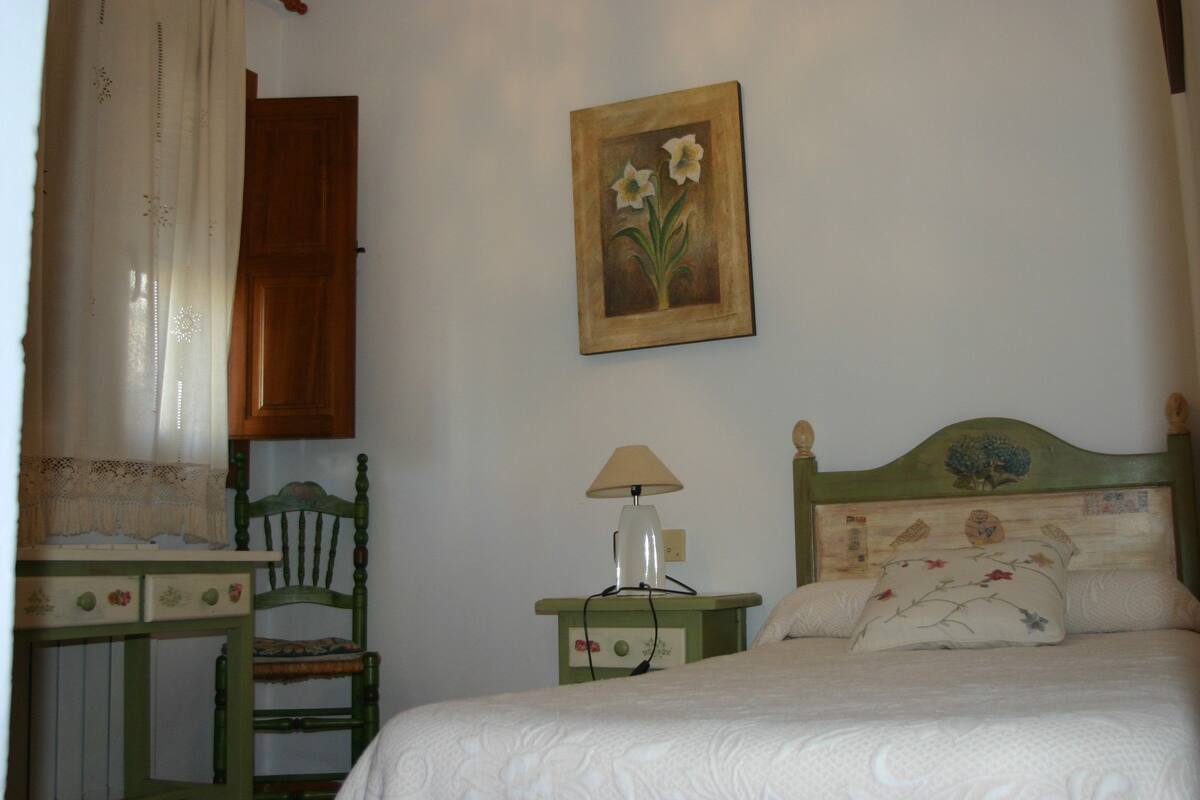 Een slaapkamer met een bed, een stoel, een nachtkastje met een lamp, een houten hoofdbord en een schilderij aan de muur.