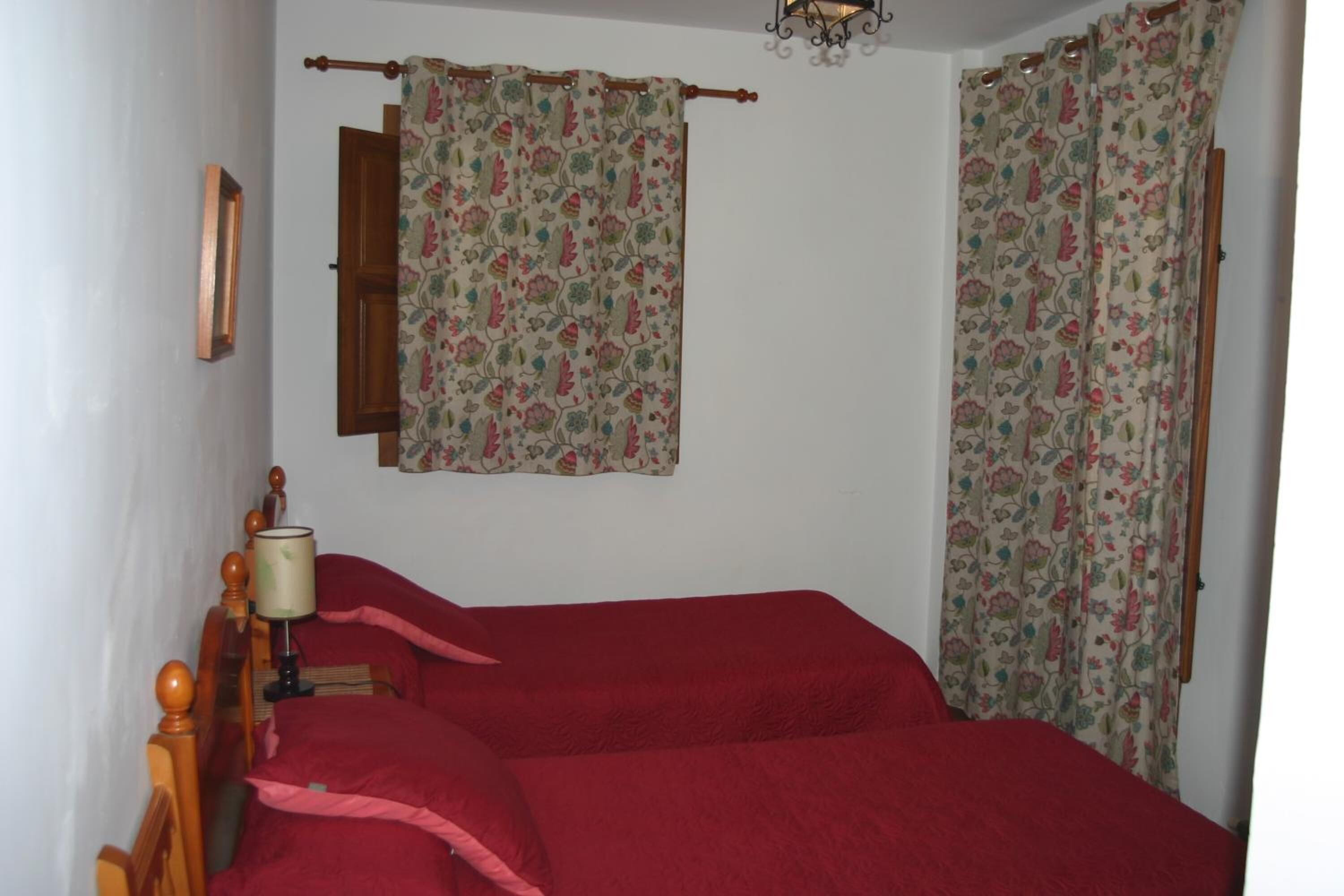 A bedroom with a wooden bed frame, red bedding, a bedside lamp, and floral curtains.