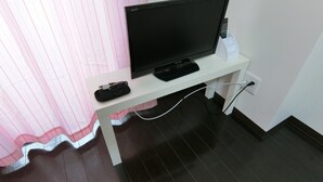 Uma televisão em um suporte branco, com um controle remoto ao lado, conectada a uma tomada na parede.