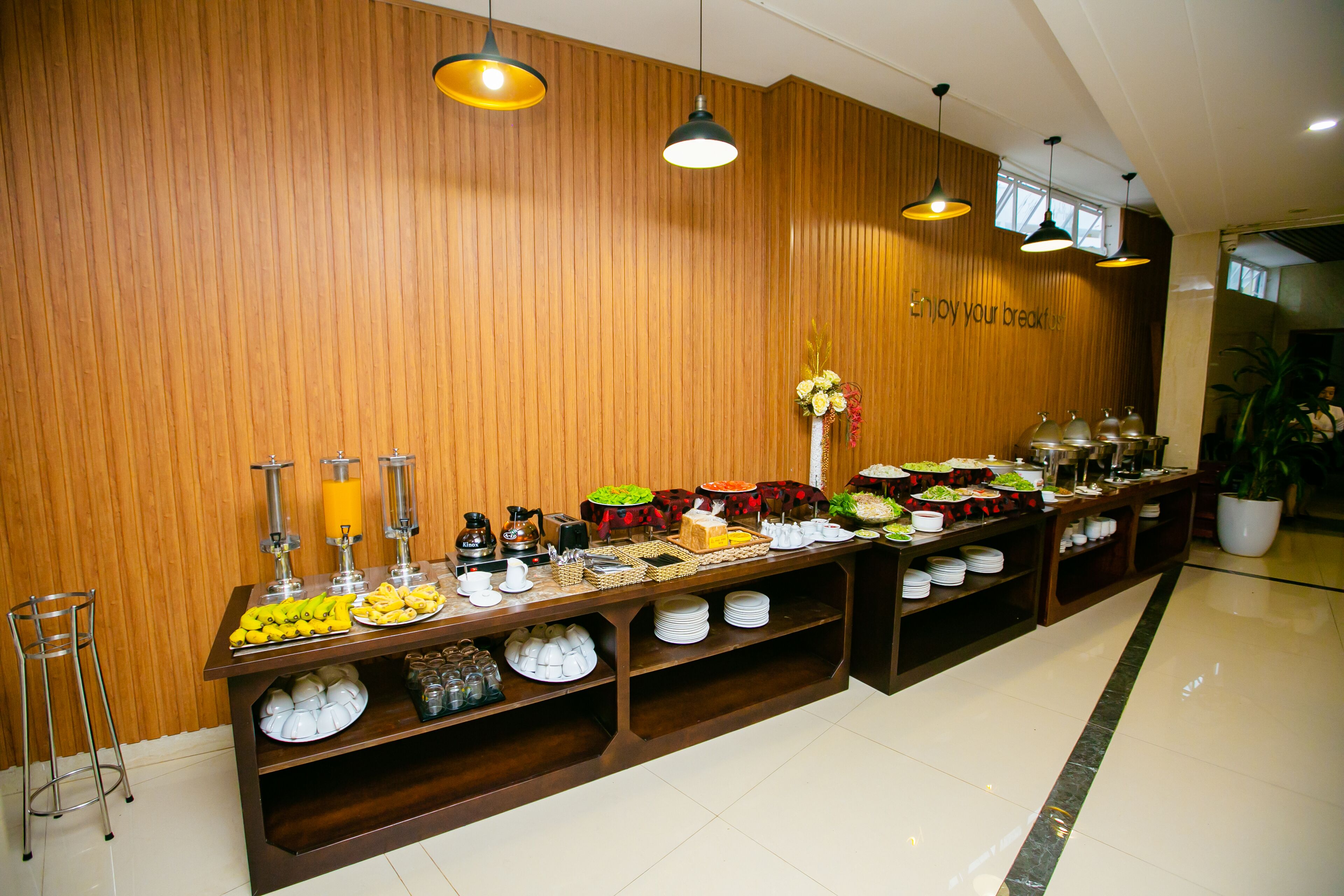 A buffet setup with various food items and beverages, wooden paneling on the wall, and hanging pendant lights.