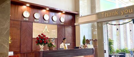 Zona de recepción de un hotel con un mostrador de madera, relojes mostrando la hora en diferentes ciudades y un cartel que dice 'Gracias' sobre el mostrador.