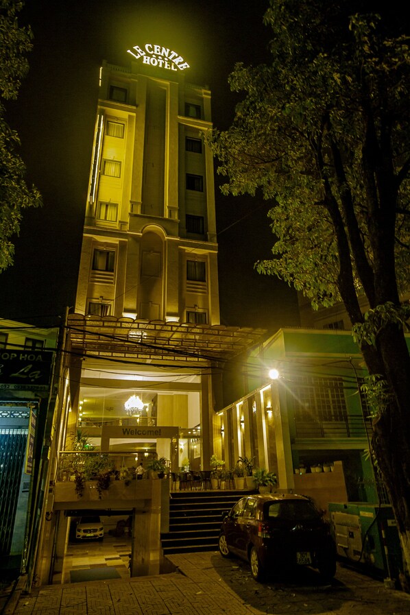 A hotel with a sign reading "Le Centre Hotel" at the top. The building has multiple floors with visible windows and balconies. There are potted plants on the ground floor and a car parked in front.