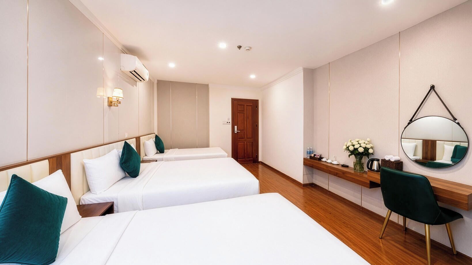 A hotel room with two beds, a wooden floor, a desk with a chair, a mirror, and a door.