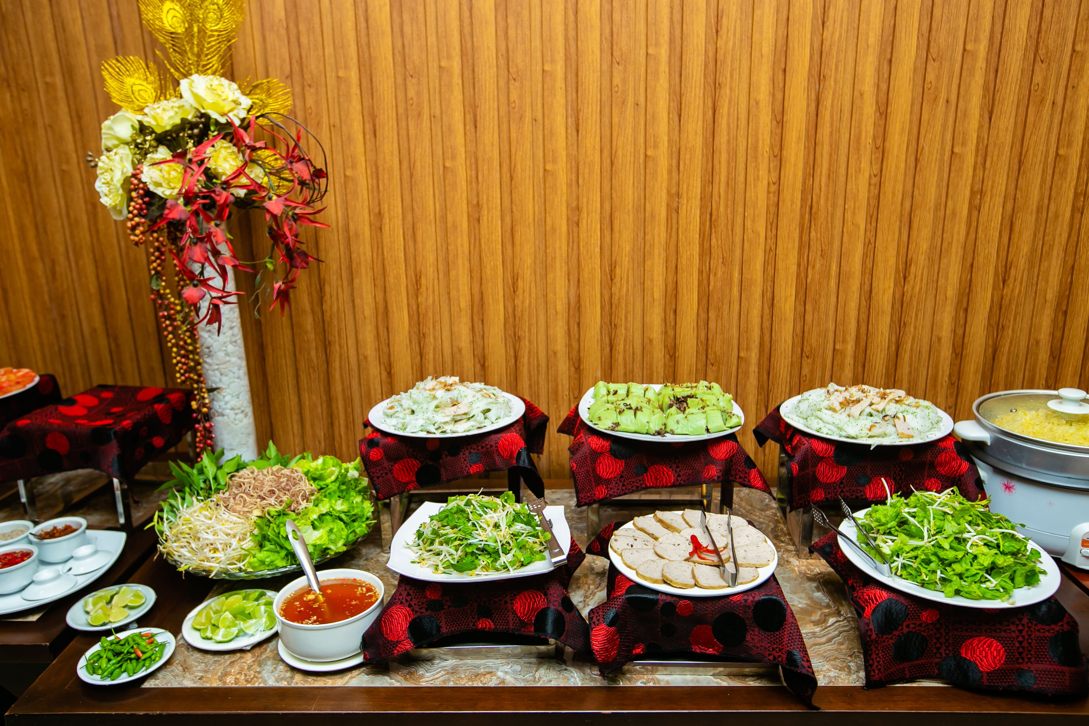 Une table de buffet proposant divers plats, notamment des nouilles, des salades et un grand plat de soupe, le tout disposé devant un décor en bois.