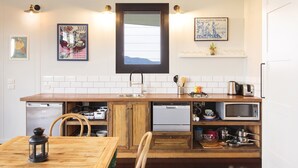 A compact kitchen with wooden cabinetry, a built-in dishwasher, a microwave, and a sink. There are framed pictures on the wall, a window above the sink, and a small dining table with chairs. - “A Secret place to slow down” (Gloucester)