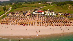 An aerial view of a beach with numerous umbrellas and people, a parking area, and buildings in the background. - Hotel Kanali (Preveza)