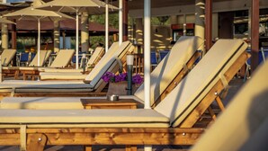 A row of wooden lounge chairs with beige cushions, arranged in a sunlit outdoor area with umbrellas. - Hotel Kanali (Preveza)