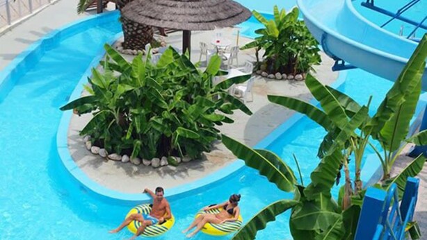 A swimming pool with two people on inflatable rings, surrounded by lush greenery and a thatched-roof structure. - Hotel Kanali (Preveza)