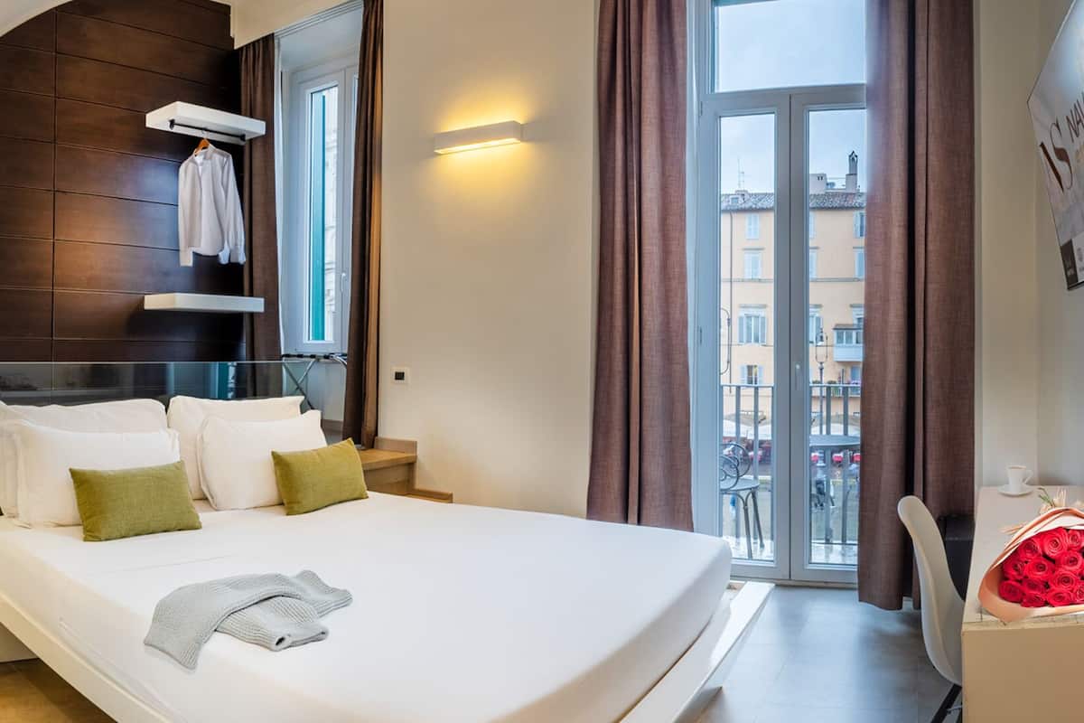 A modern hotel room with a large bed, a desk, and a view of the city through large windows.