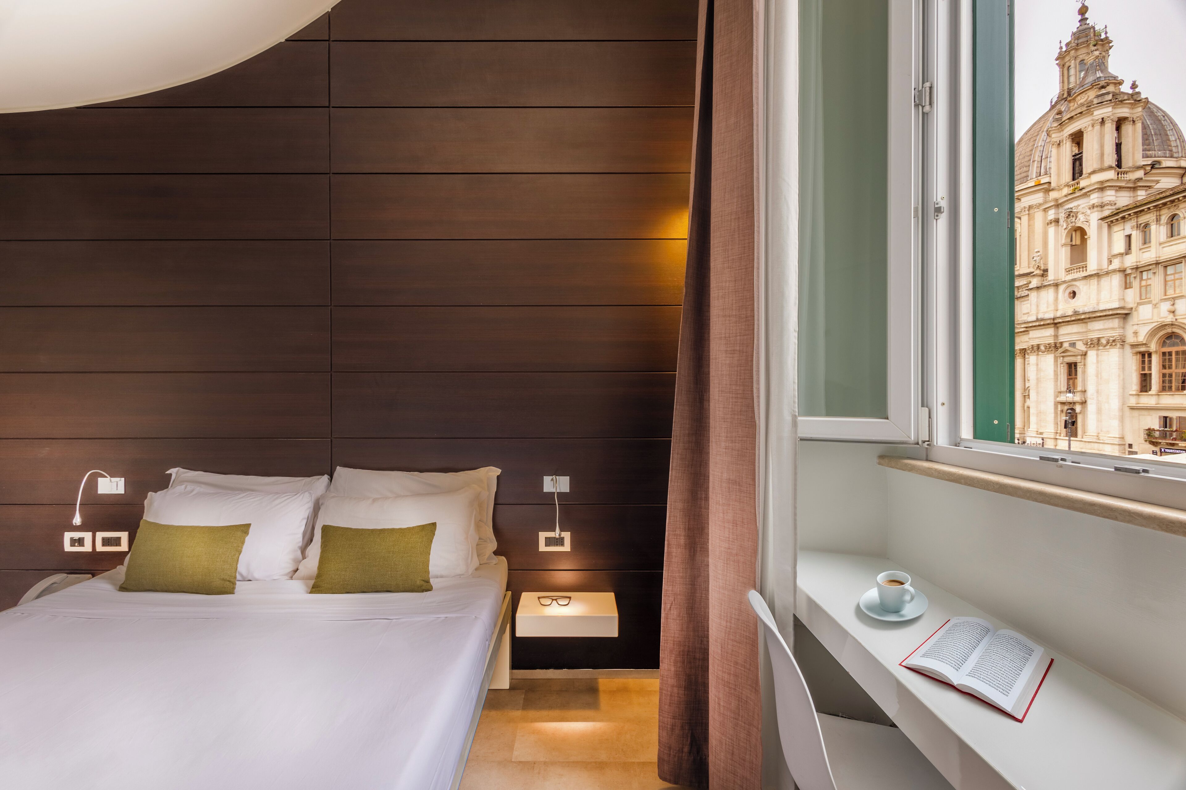 A modern hotel room with a large bed, bedside lamps, a nightstand, a window with a view of a historic building, and a small table with a book and cup.