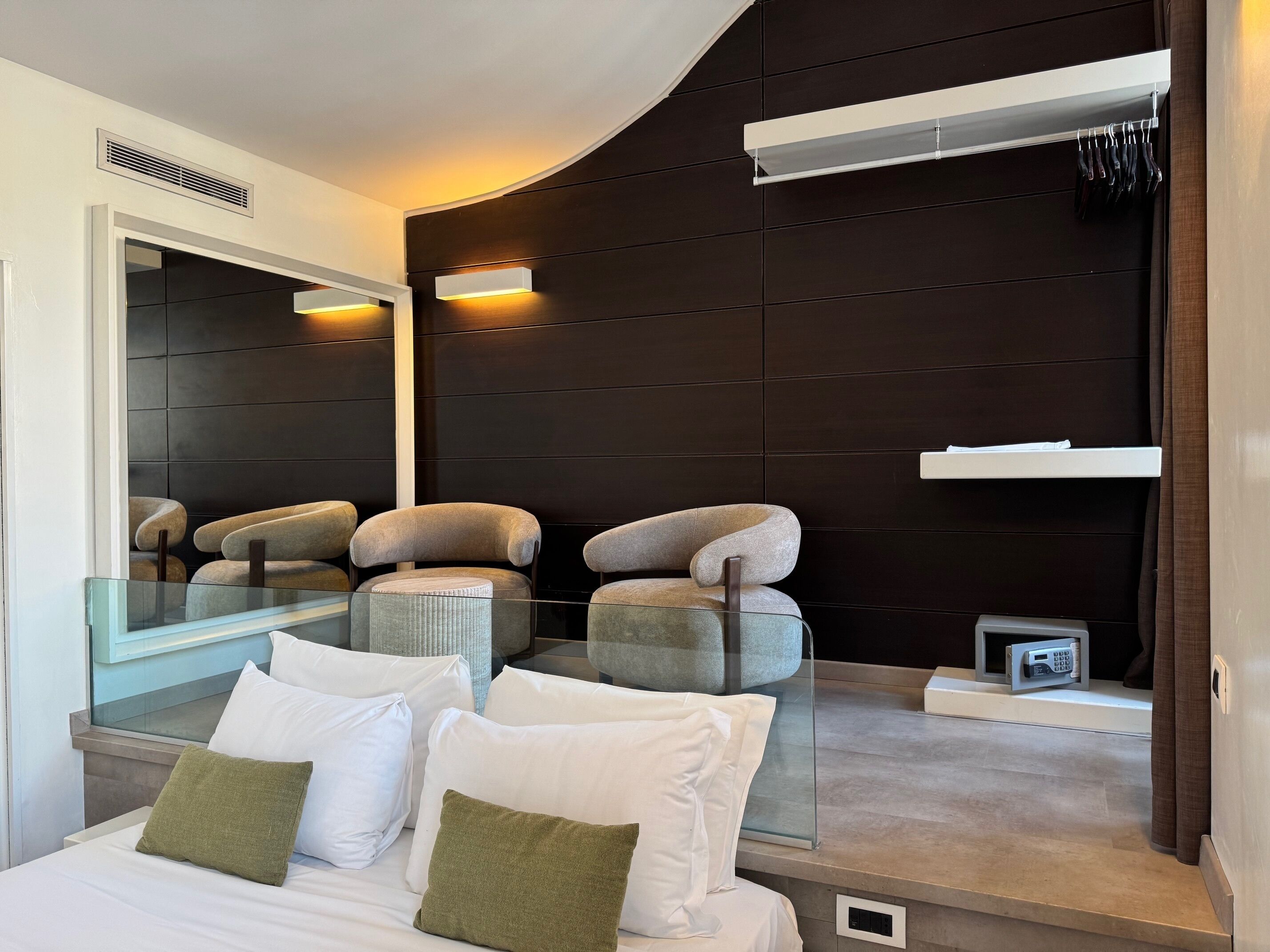 A modern hotel room with a curved dark wall, a large mirror, a glass table, and two cushioned chairs.