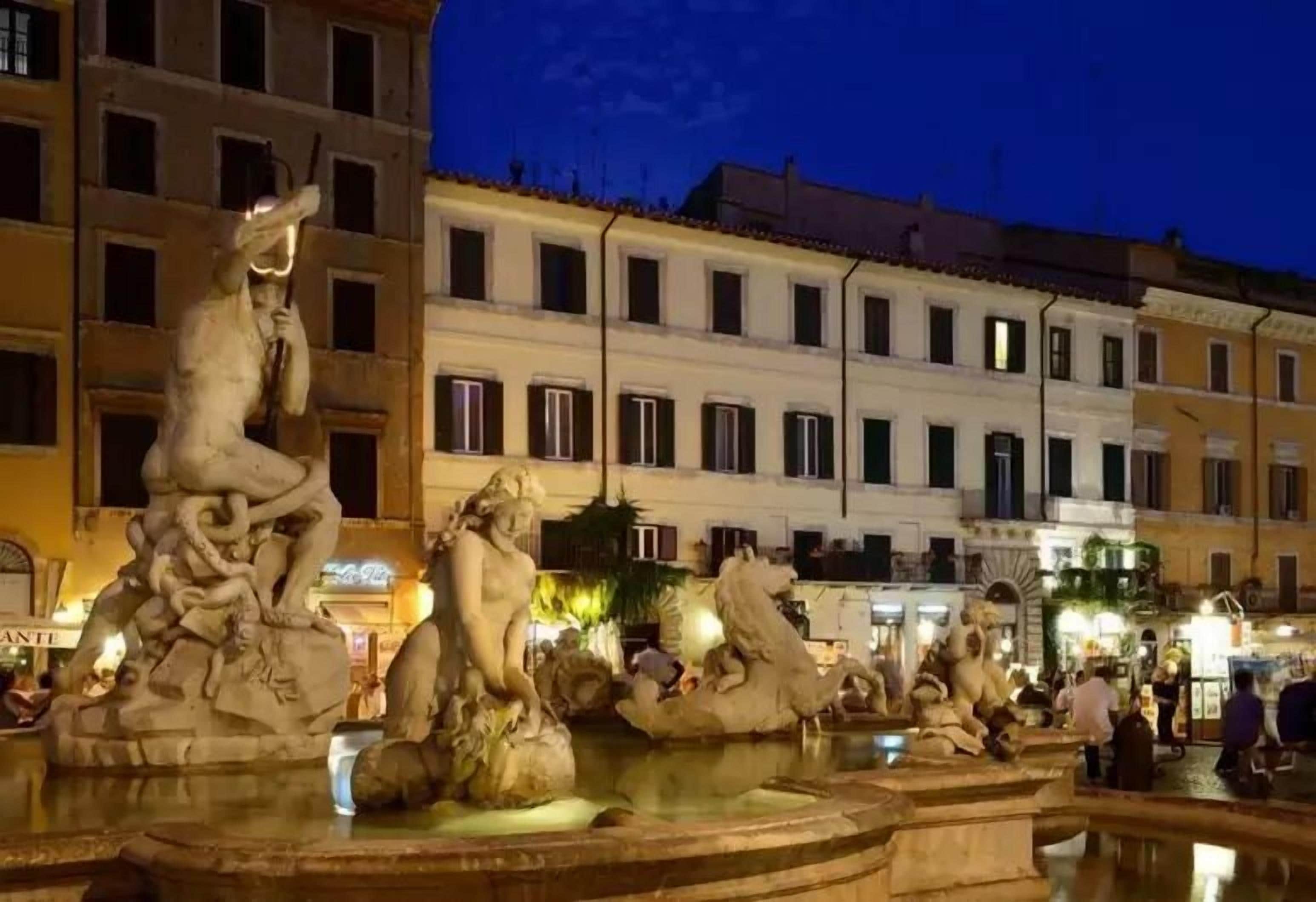A public square with a classical fountain featuring multiple statues, surrounded by buildings with uniform windows and balconies.