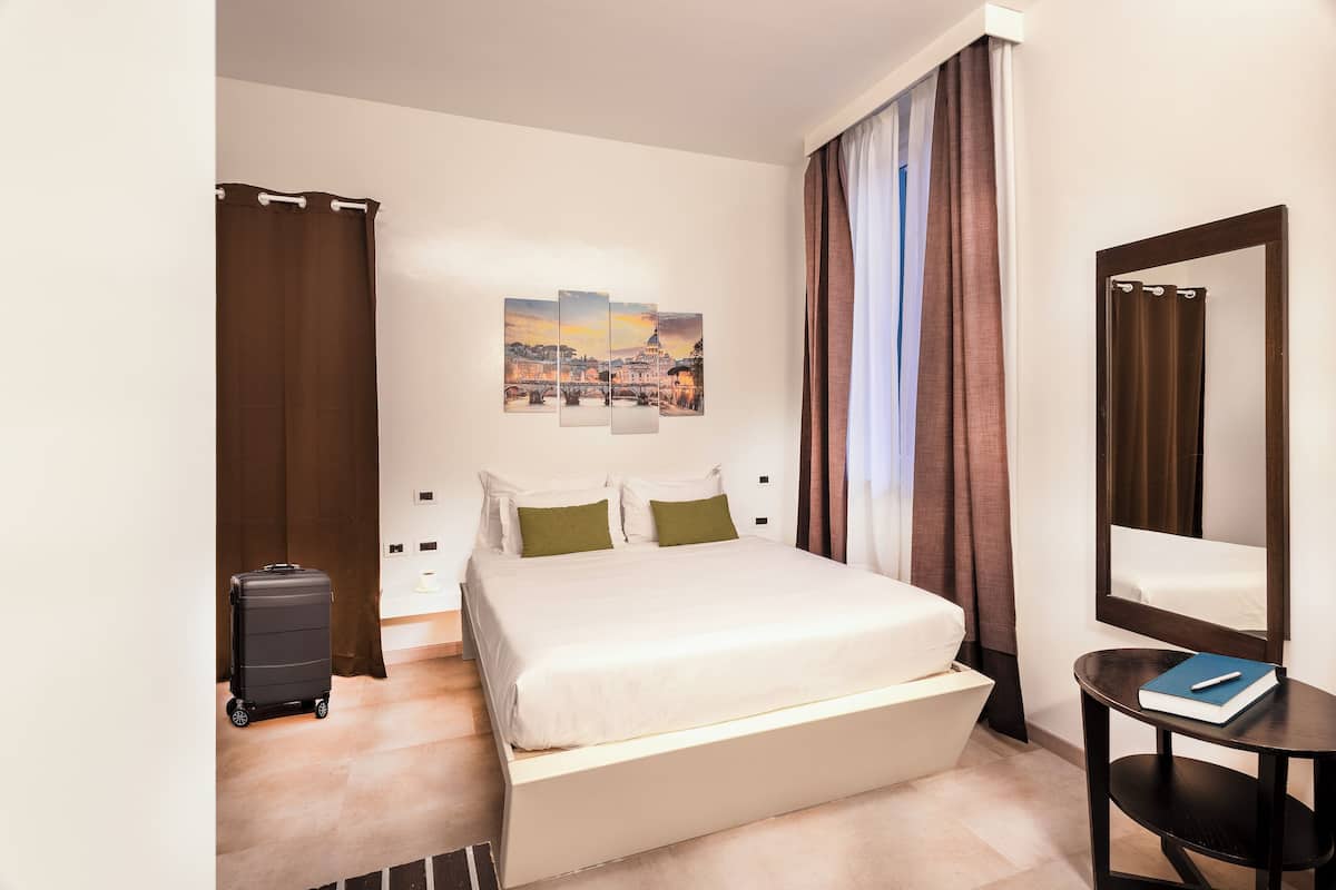 A hotel room with a bed, a small table with a book, a suitcase, and a painting of a cityscape on the wall.