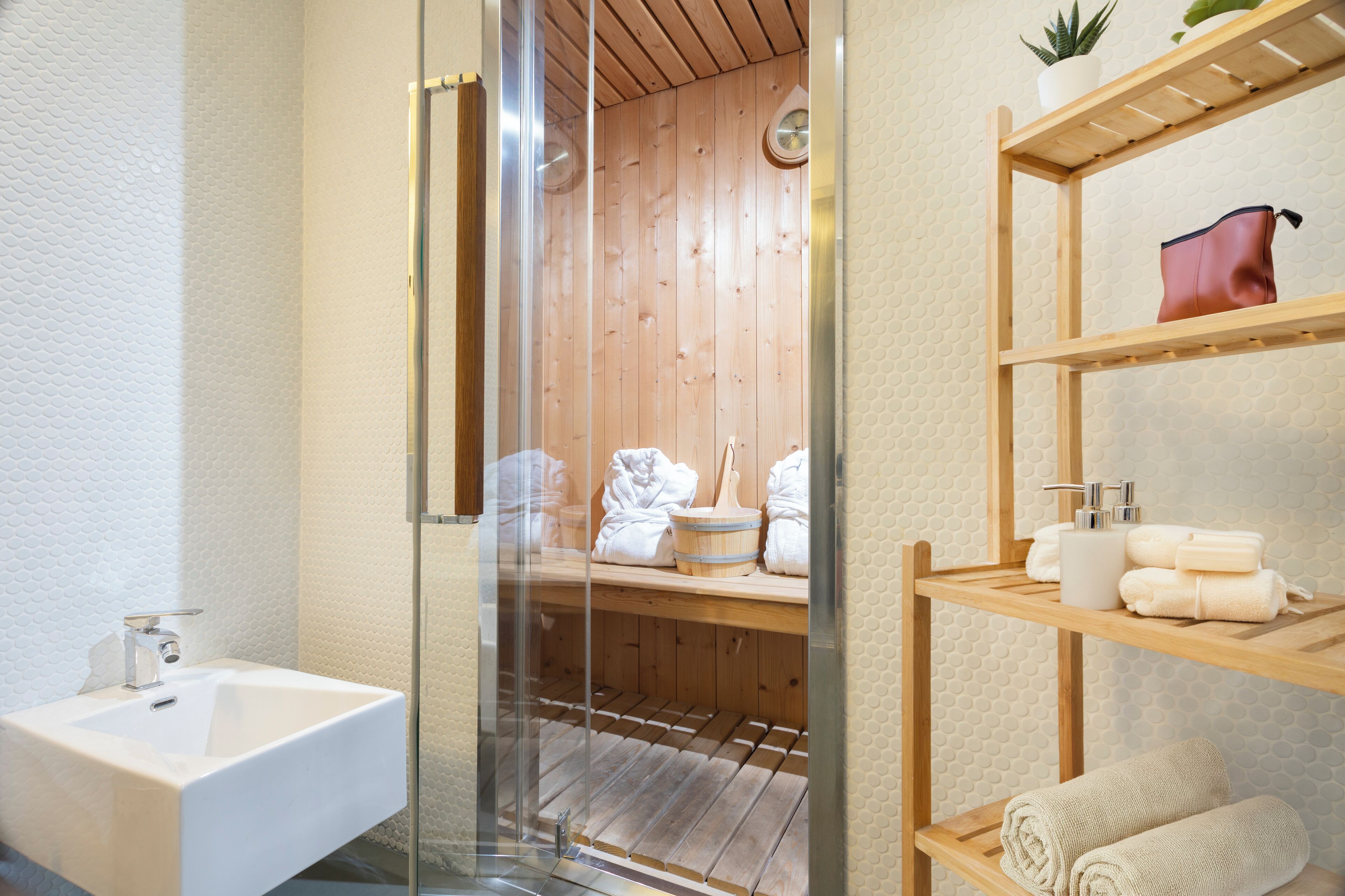 Une salle de bain moderne avec un sauna à parois de verre, une étagère en bois où sont posées des serviettes et un lavabo blanc.