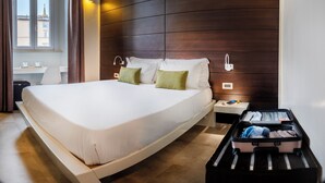 Camera d'albergo con un letto grande, una scrivania con due tazze, una valigia e vista sugli edifici attraverso una finestra.