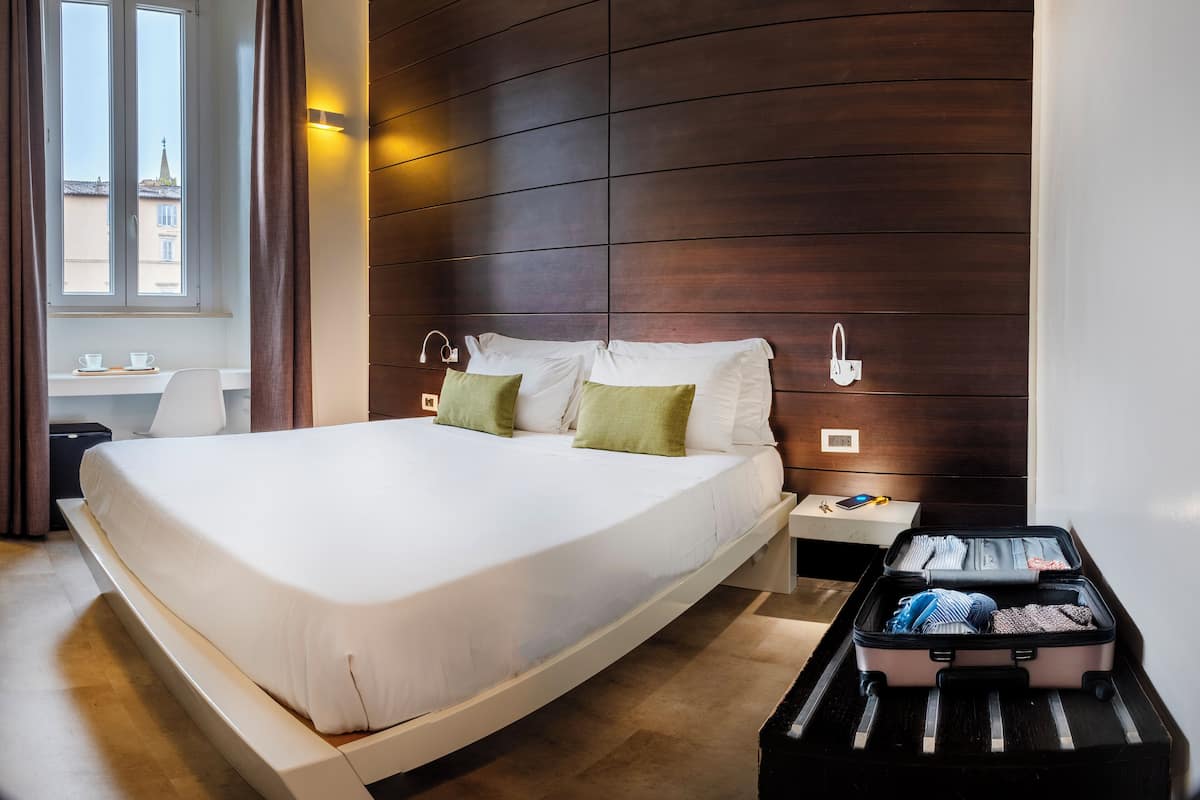 A hotel room with a large bed, a desk with two cups, a suitcase, and a view of buildings through a window.