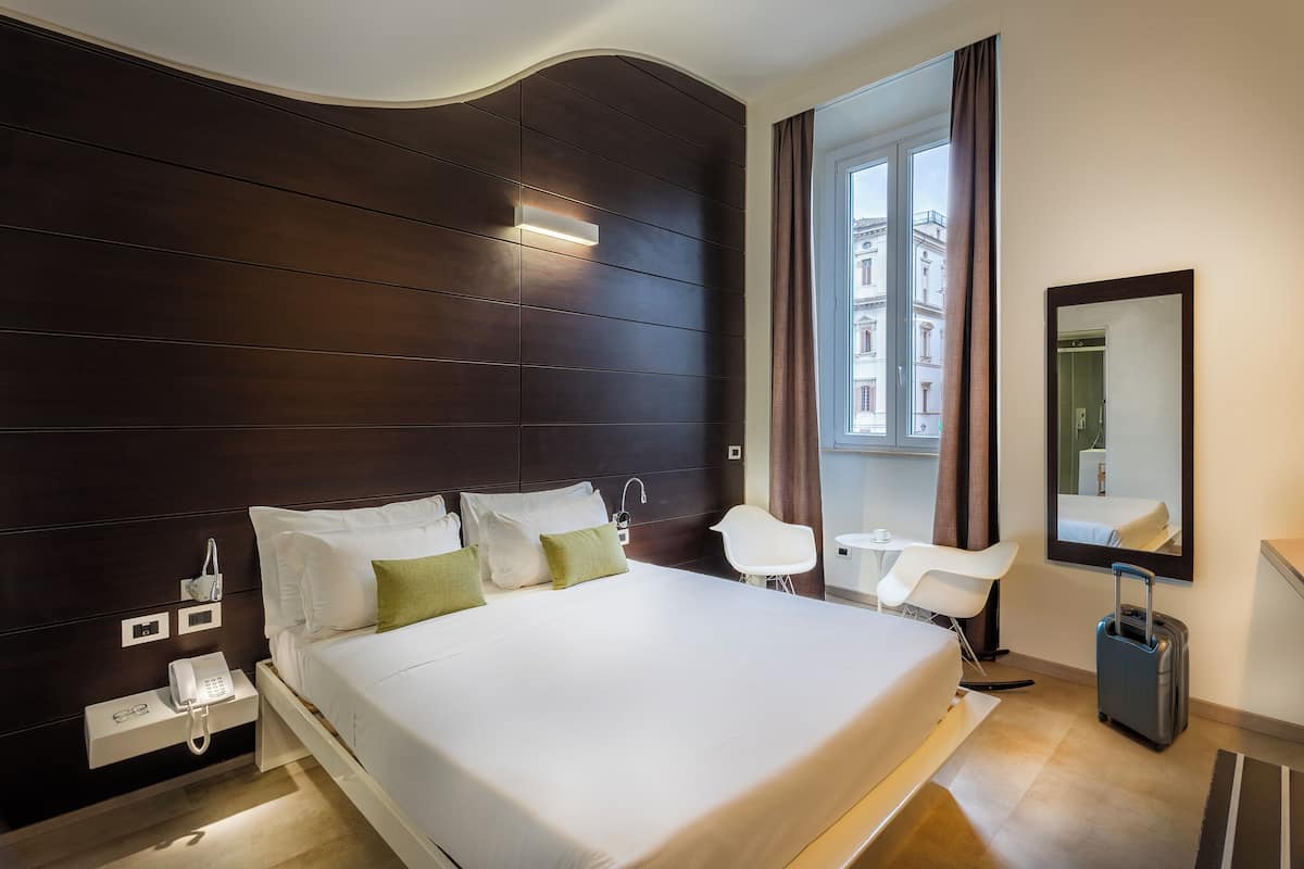 A modern hotel room with a large bed, bedside table with a phone, a small desk, and a chair.