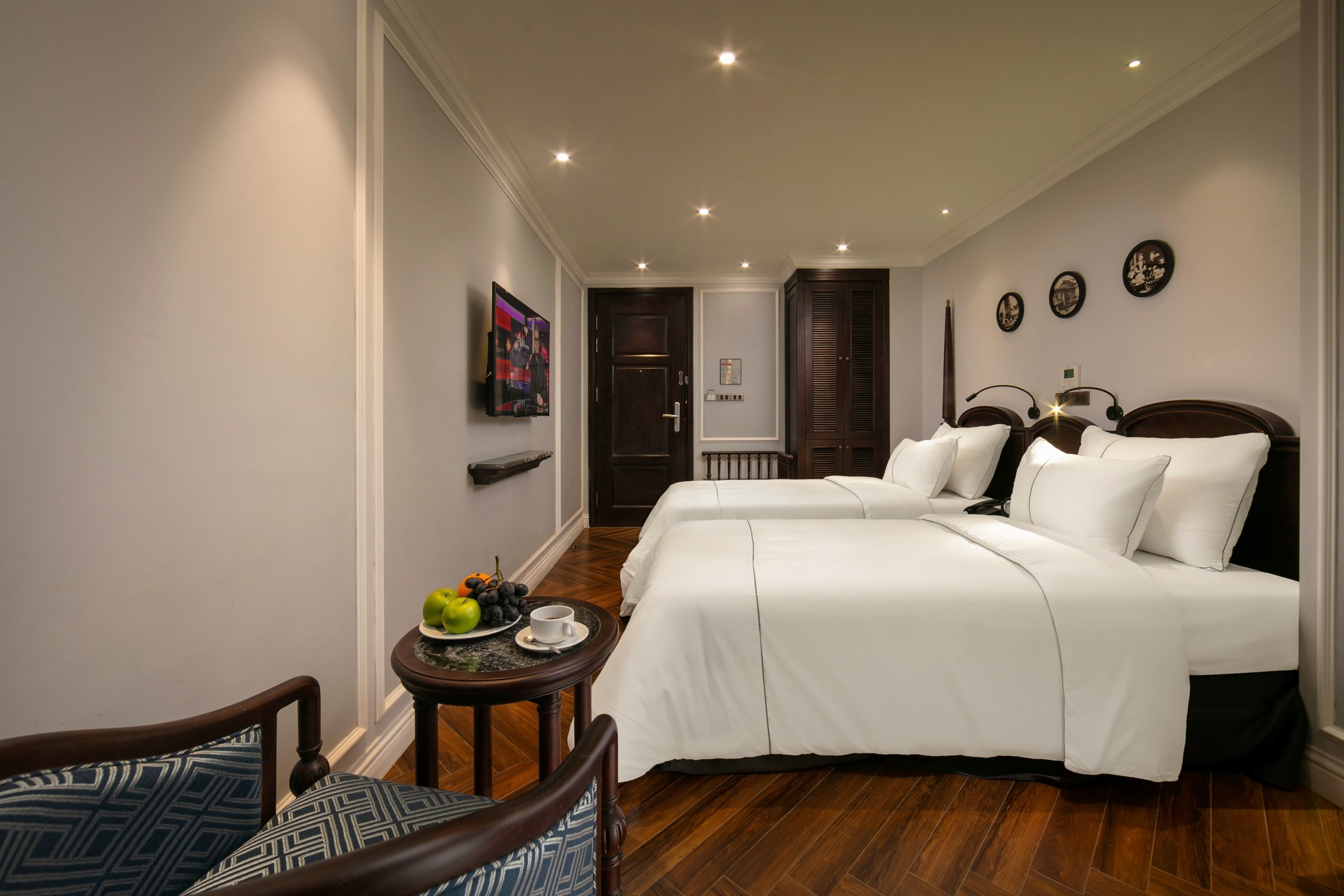A hotel room with two beds, a small table with fruit and a cup, and a chair with patterned cushions.