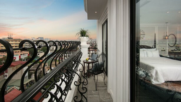 Un balcon donnant sur le paysage urbain, une petite table avec une chaise et un lit recouvert d’une courtepointe blanche.