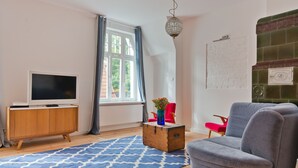 A living room with a wooden cabinet, a grey sofa, a red chair, a blue rug, and a large window with curtains. - Imperial Apartments - Winieckiego (Sopot)