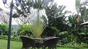 A garden with a wicker table and chairs, surrounded by various tropical plants and a stone wall. - Sherbourne Lodge (Kitwe)