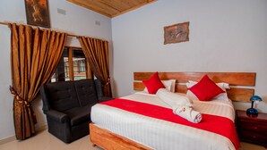 A bedroom with a wooden bed, red pillows, a black armchair, and a wooden nightstand with a lamp. - Sherbourne Lodge (Kitwe)