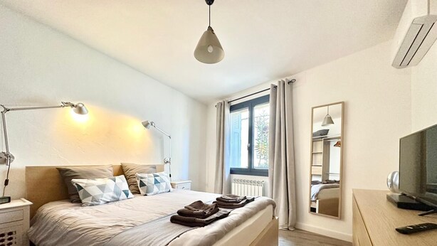 A modern bedroom with a large bed, bedside lamps, a TV, and a window with curtains. - Home 12 "les Vignes" (Châteauneuf-du-Pape)