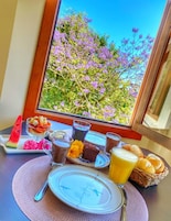 Uma mesa de café da manhã com diversos alimentos e bebidas, um prato com flores cor de rosa e vista para flores roxas através de uma janela.