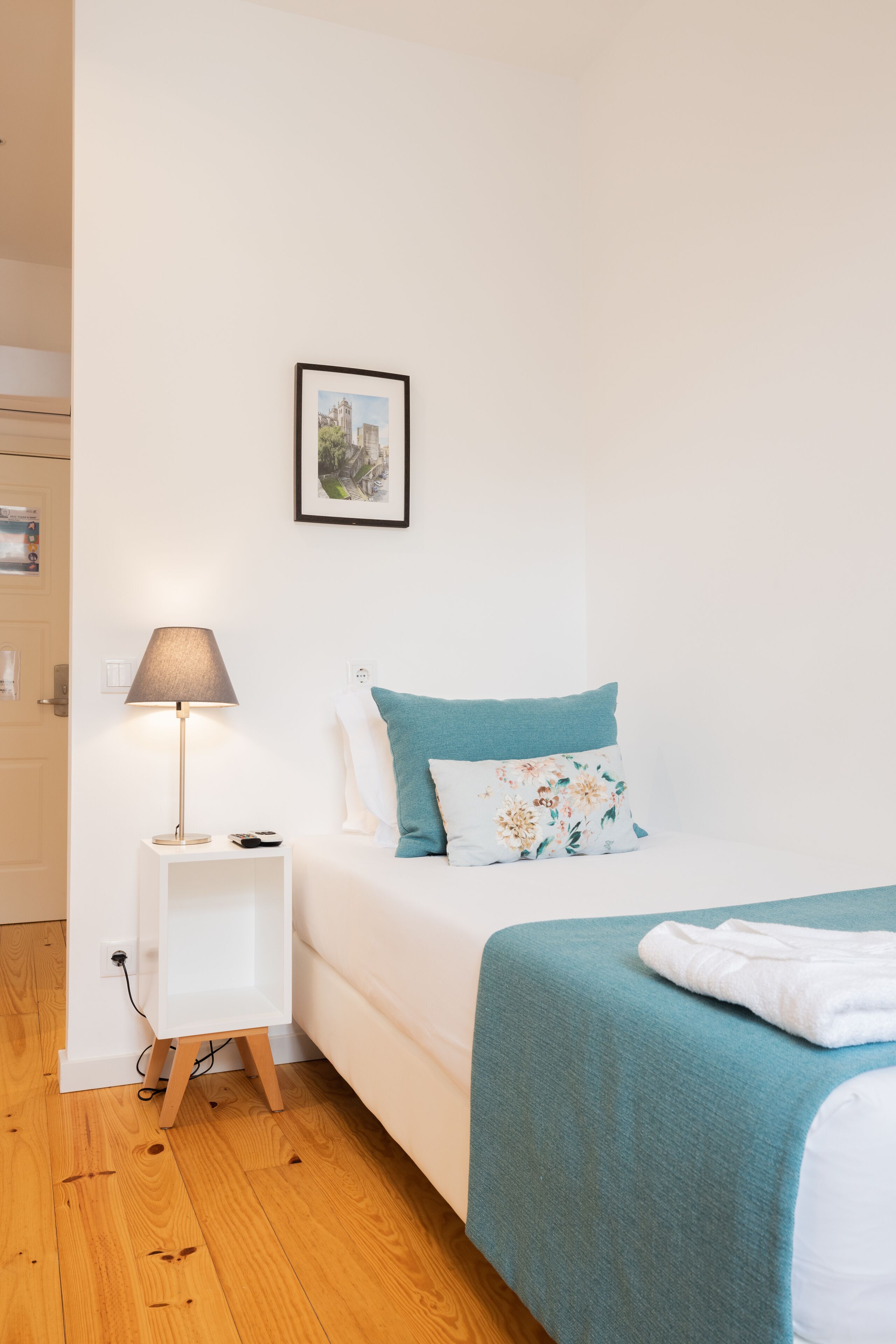 A neatly arranged bedroom with a bed, a bedside table with a lamp, a small shelf, and a framed picture on the wall.