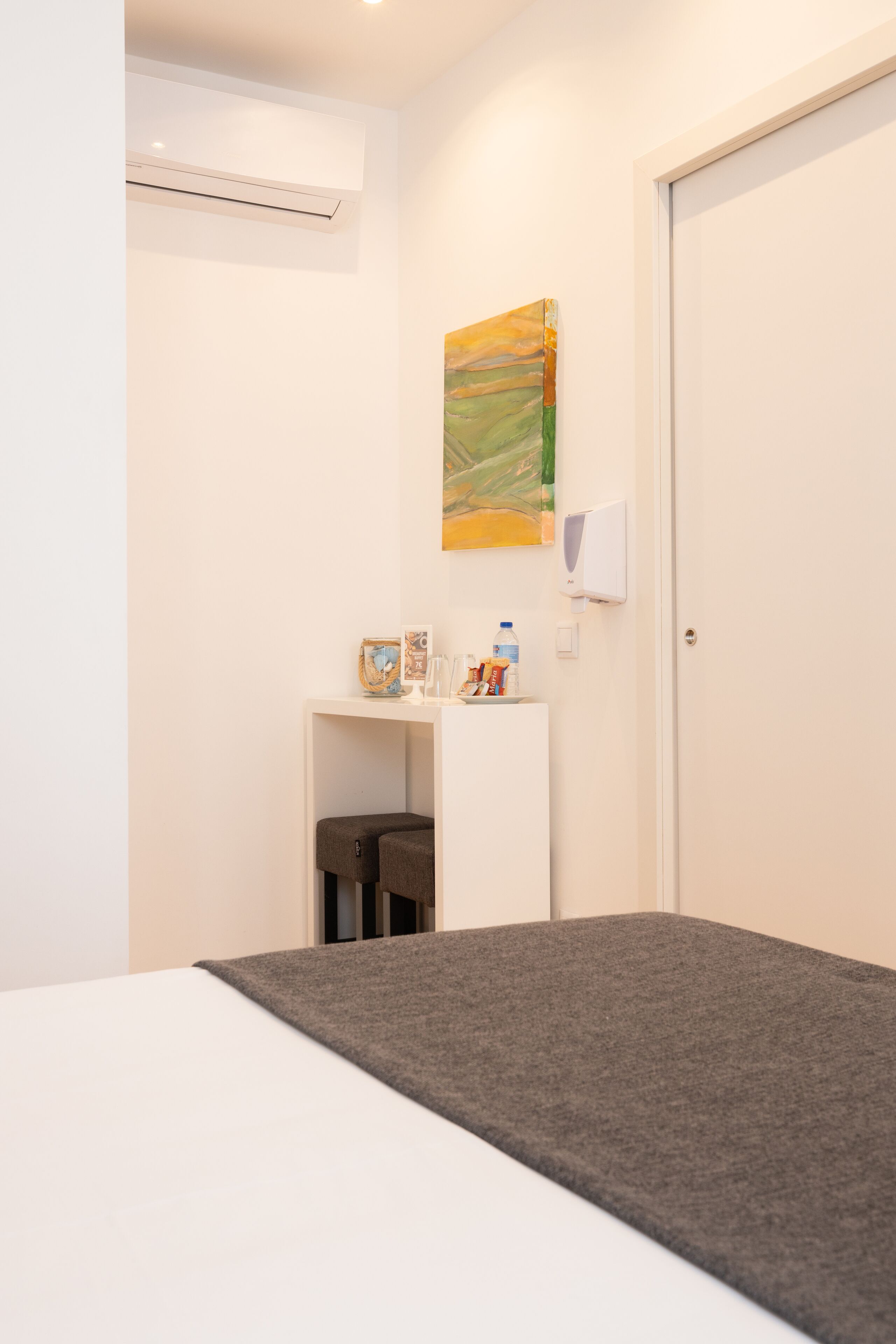 A hotel room with a bed, a small desk with a water bottle, a painting on the wall, and a door.