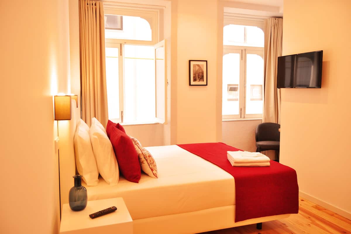 A hotel room with a bed, a TV mounted on the wall, a chair, and a window with curtains.